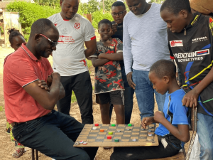 Hon. Binwell Chansa Mpundu plays a bottle-cap board game with local youth in Nkana, Kitwe, as community members look on